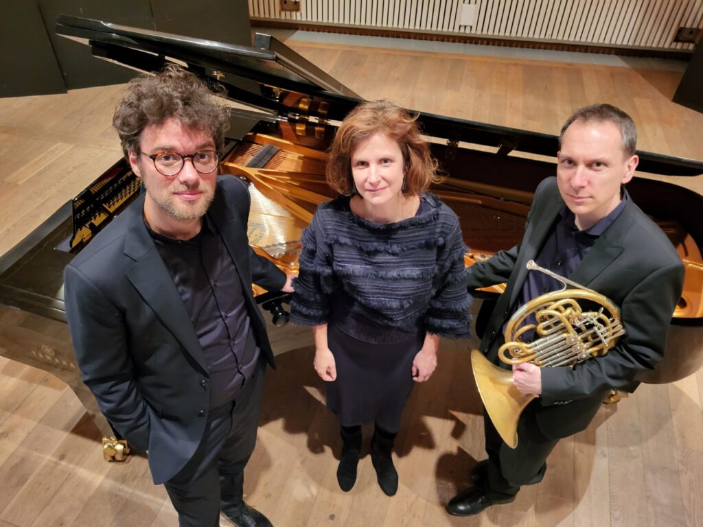 Drie muzikanten (man, vrouw, man) poseren in donkere kleding voor een vleugel piano, de man rechts houdt een hoorn vast