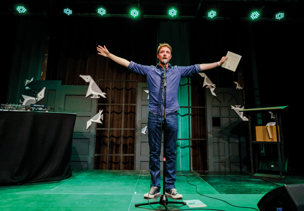 foto max greyson aan microfoon op podium, armen uitgestrekt papieren in een hand